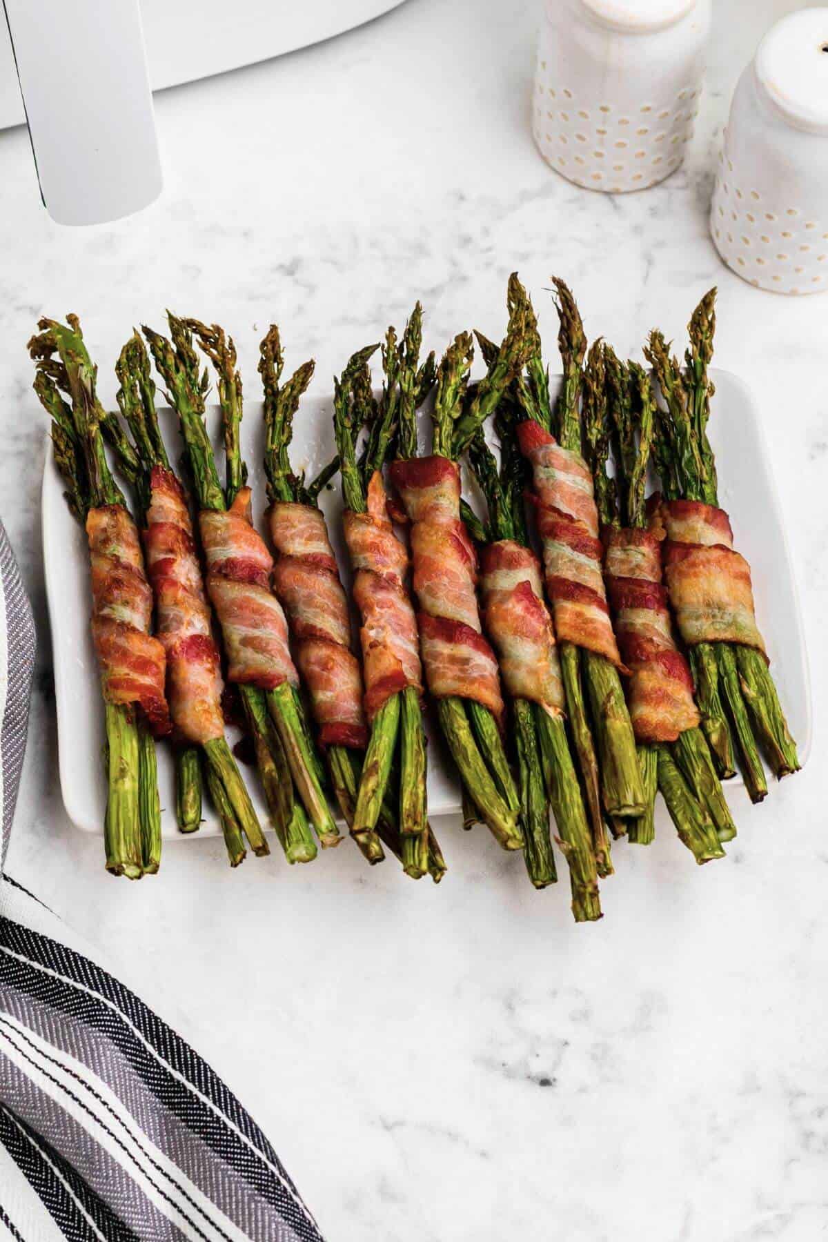 Crispy golden brown bacon wrapped around asparagus stalks on a white rectangle plate. 