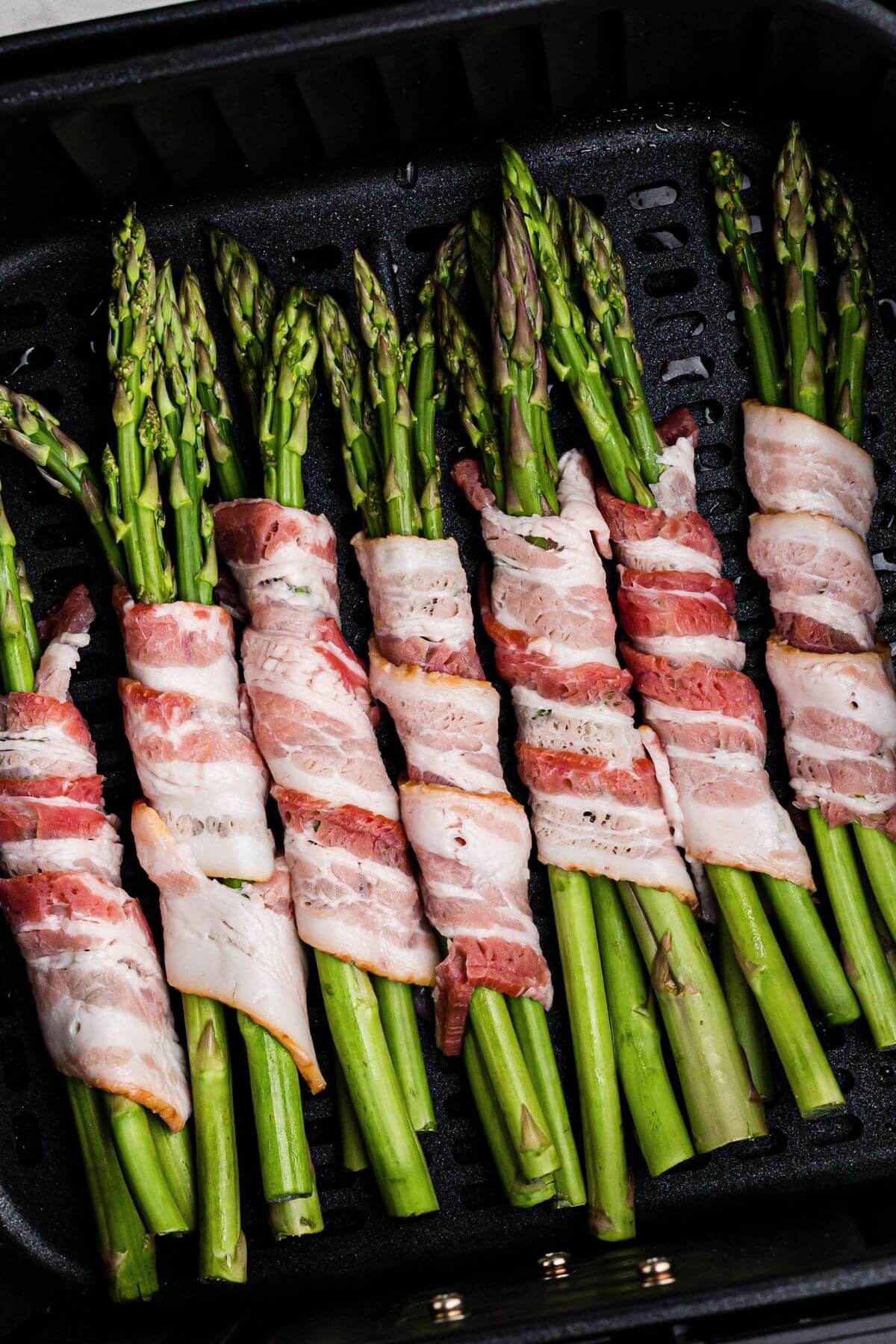Uncooked bacon wrapped around uncooked asparagus spears in the air fryer basket. 