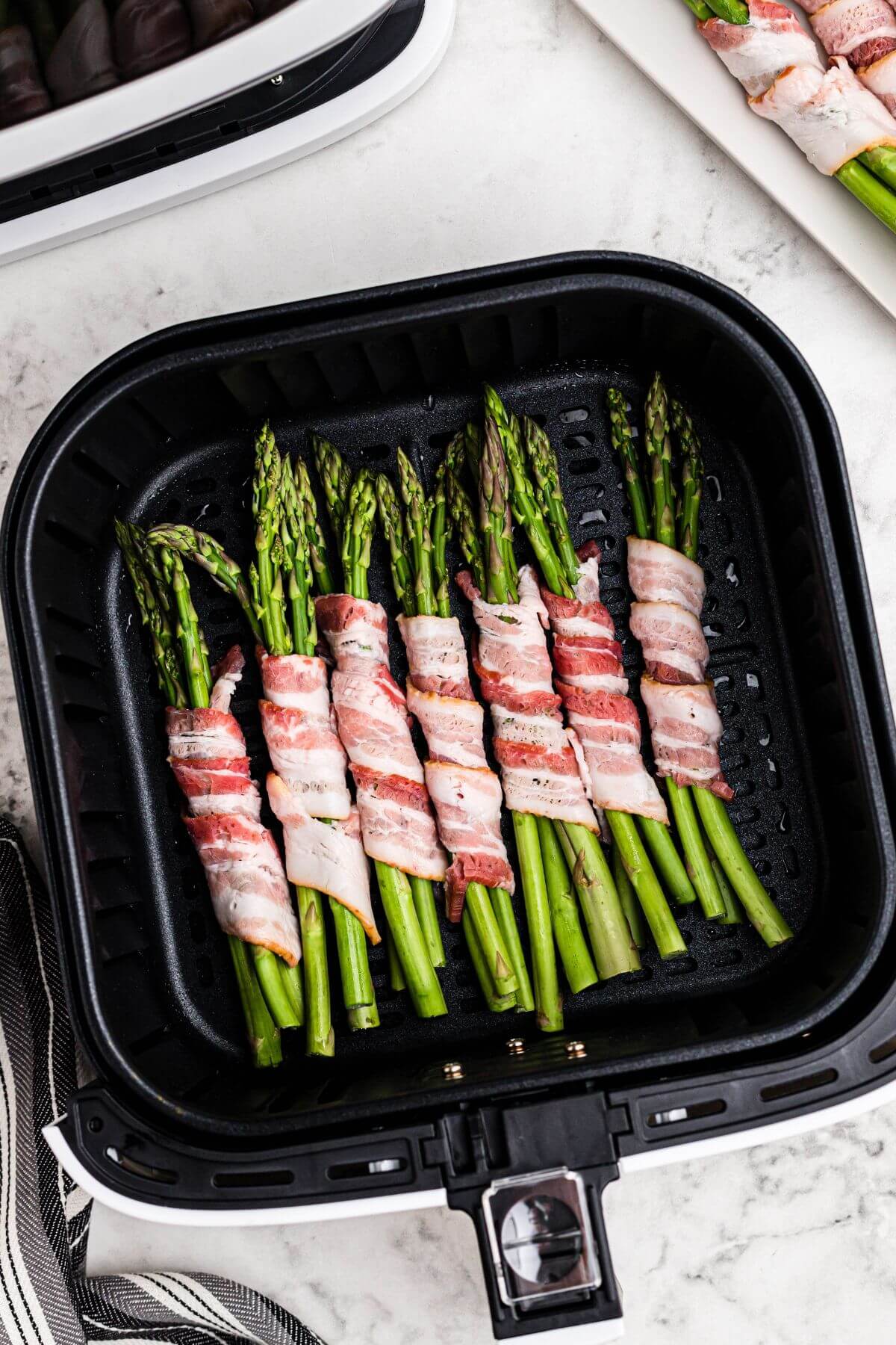 Uncooked bacon slices wrapped around asparagus spears in the air fryer basket. 