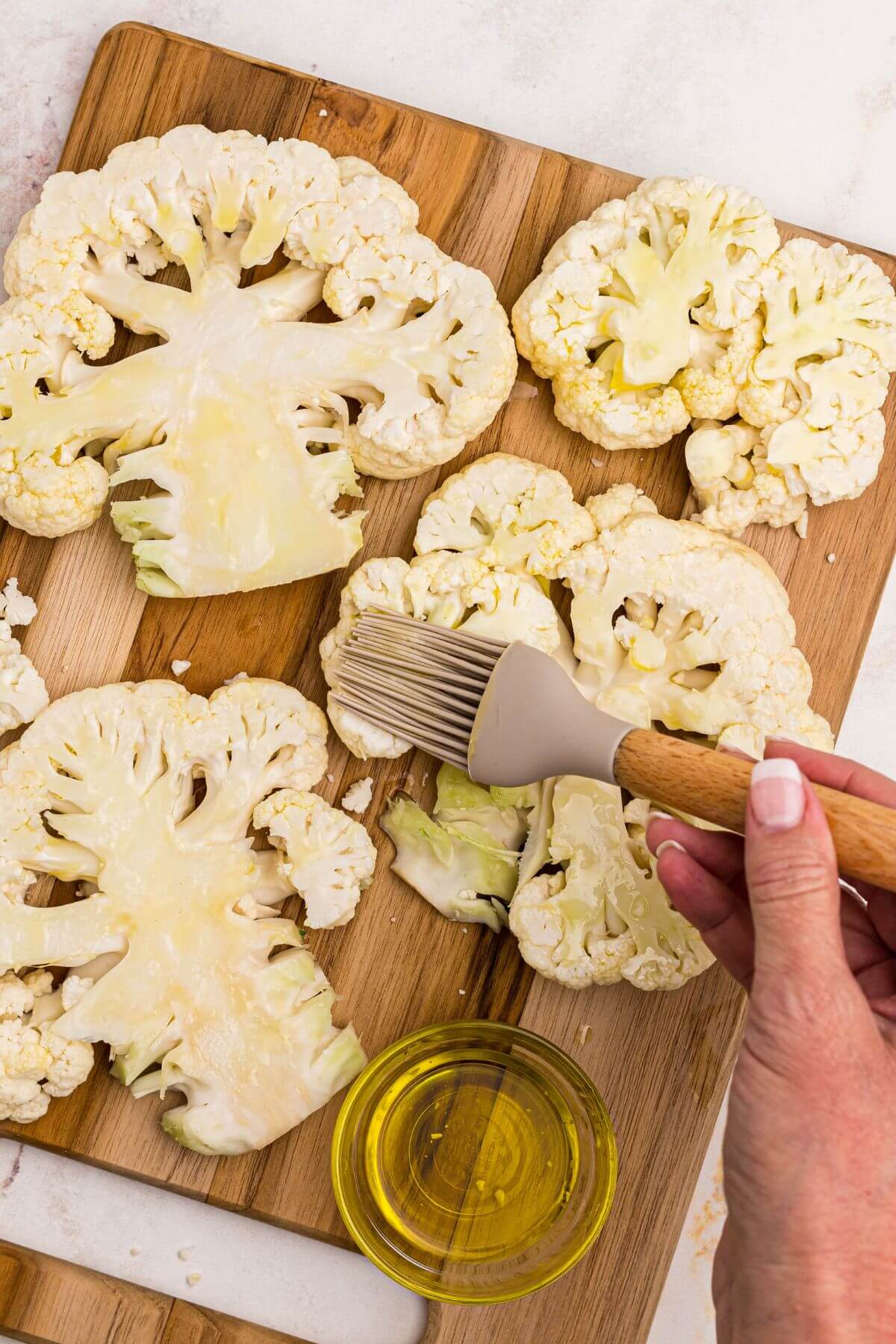 Uncooked cauliflower steaks on a wooden cutting board being brushed with olive oil.