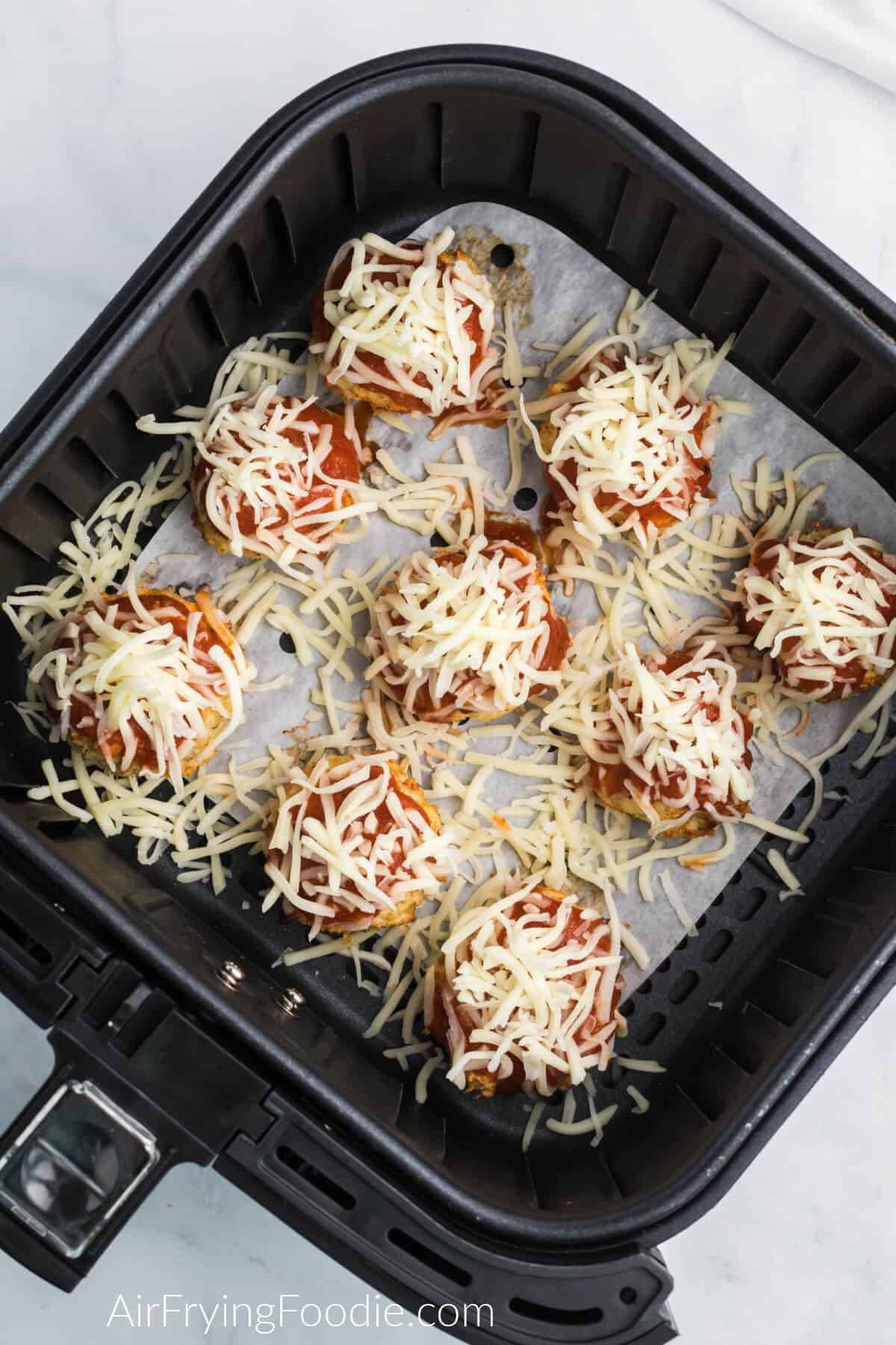 Chicken meatballs in air fryer basket on top of parchment paper topped with marinara sauce, parmesan cheese, and mozzarella cheese. 