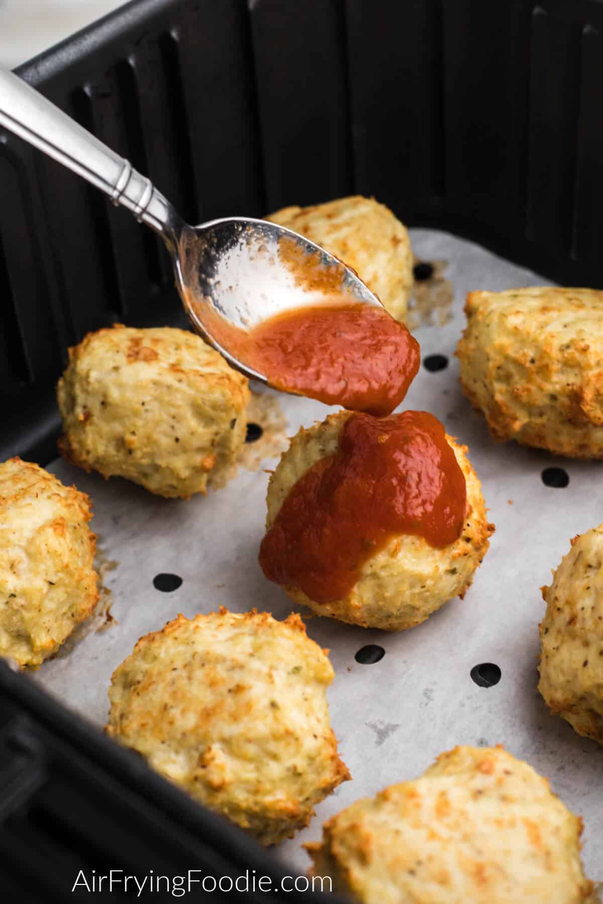 Chicken meatballs in air fryer basket on top of parchment paper, being topped with marinara sauce. 