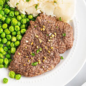 Juicy cube steak on a white plate served with green peas and mashed potatoes.