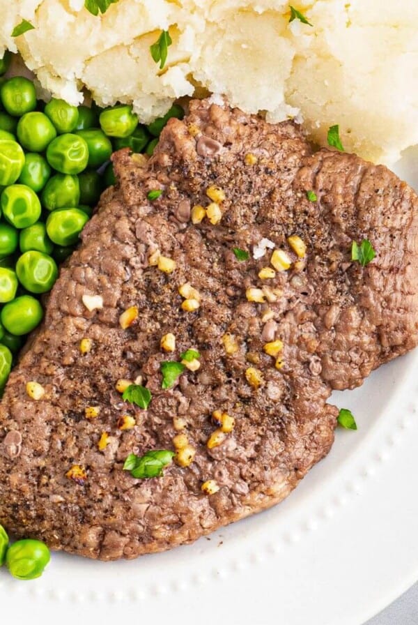 Juicy cube steak on a white plate served with green peas and mashed potatoes.