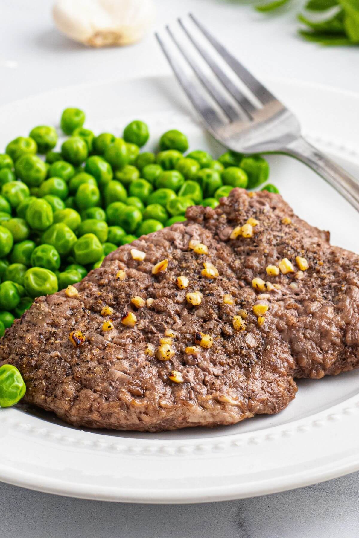 Juicy cube steak seasoned and served on a small white plate with green peas. 
