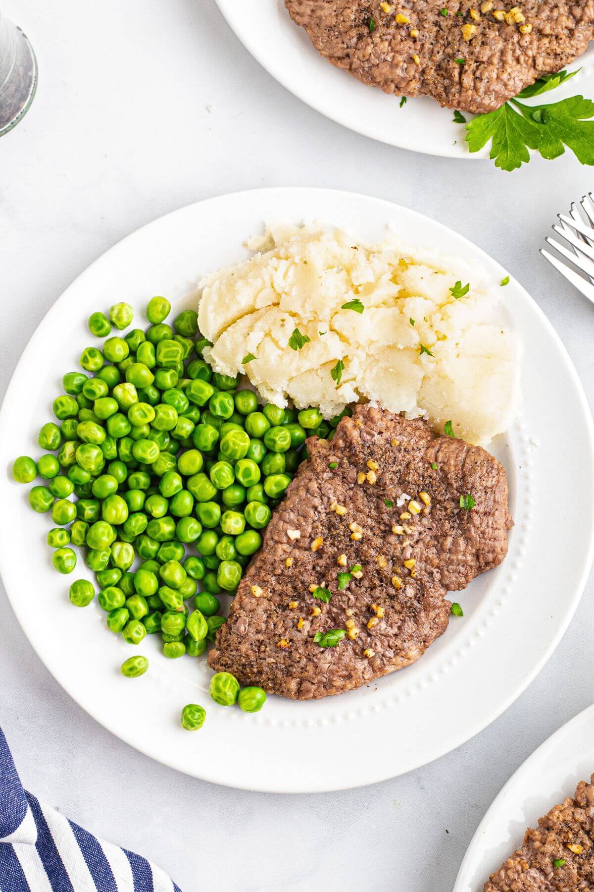 Juicy seasoned cube steak served on a white plate with green peas and mashed potatoes. 