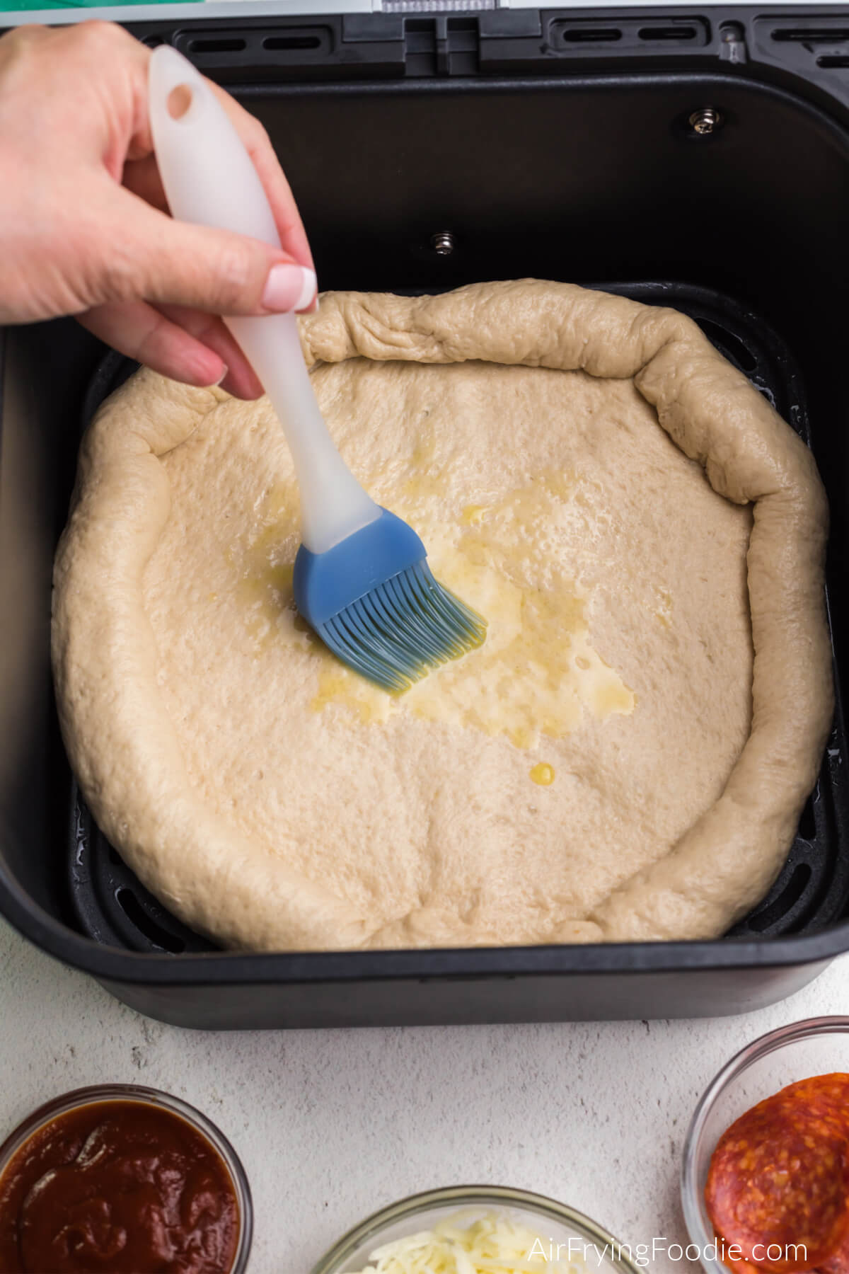 Pizza dough in the basket of the air fryer being brushed with olive oil. 