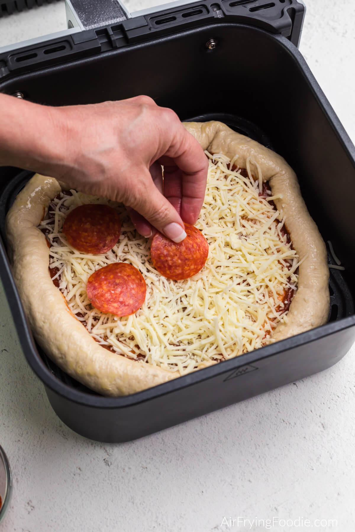 Pepperoni being placed onto a pizza in air fryer basket.