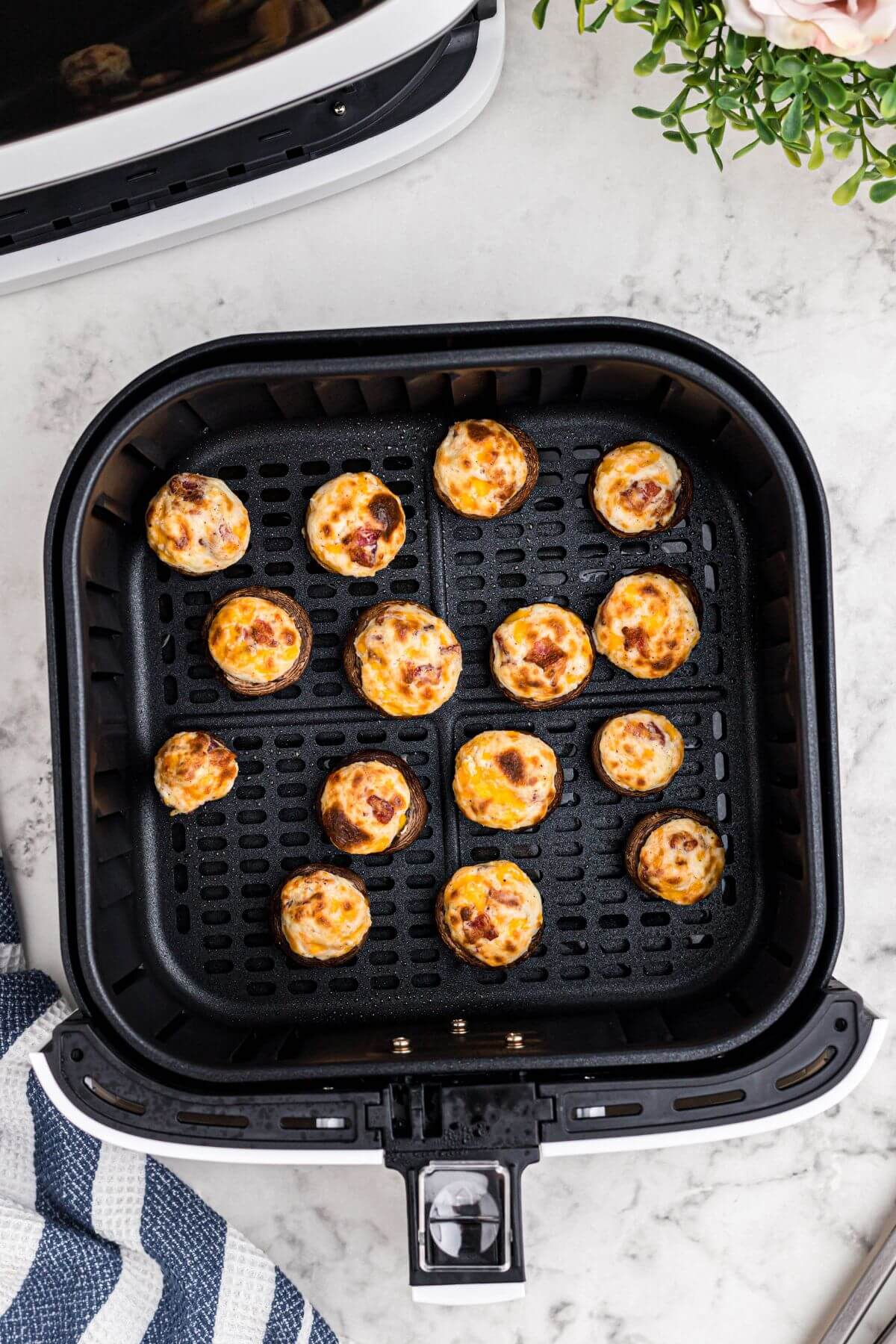 Golden brown mushrooms stuffed with cream cheese, cheddar cheese, bacon crumbles, in the air fryer basket after being cooked. 