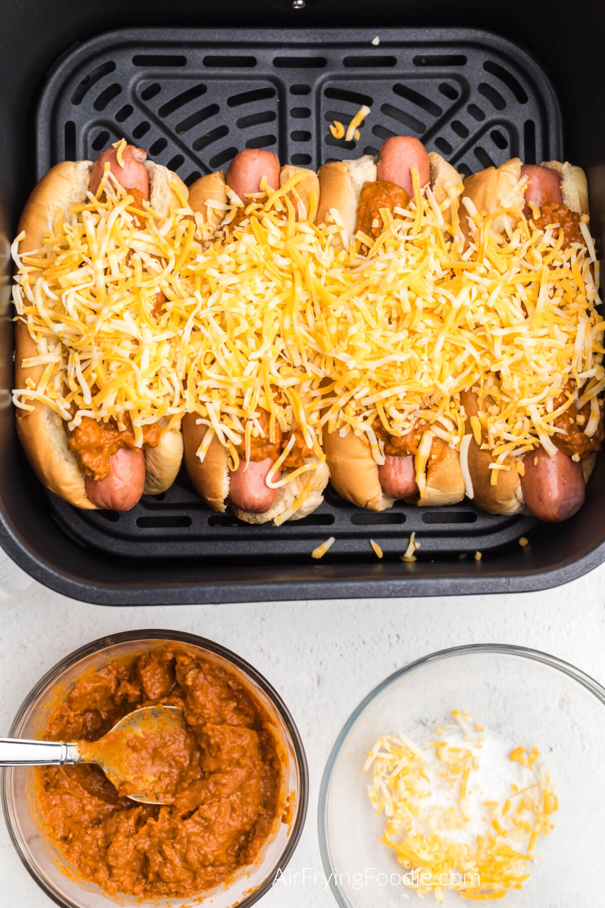 Hot dogs topped with chili and cheese in the air fryer basket, ready to be air fried. 