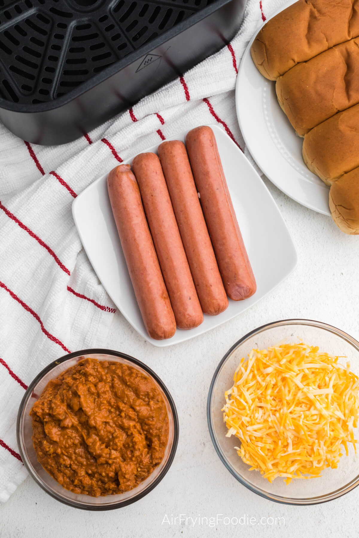Ingredients needed to make chili cheese dogs in the air fryer on a table. Hotdogs on a plate, buns on a plate, chili in a bowl, and shredded cheese in a bowl. 