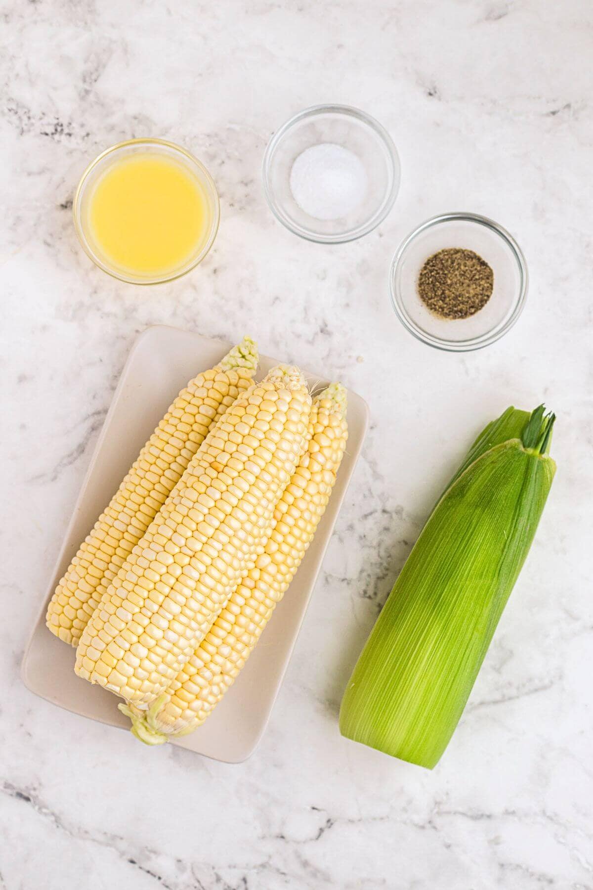 Uncooked corn on a white marble table with seasonings and melted butter. 