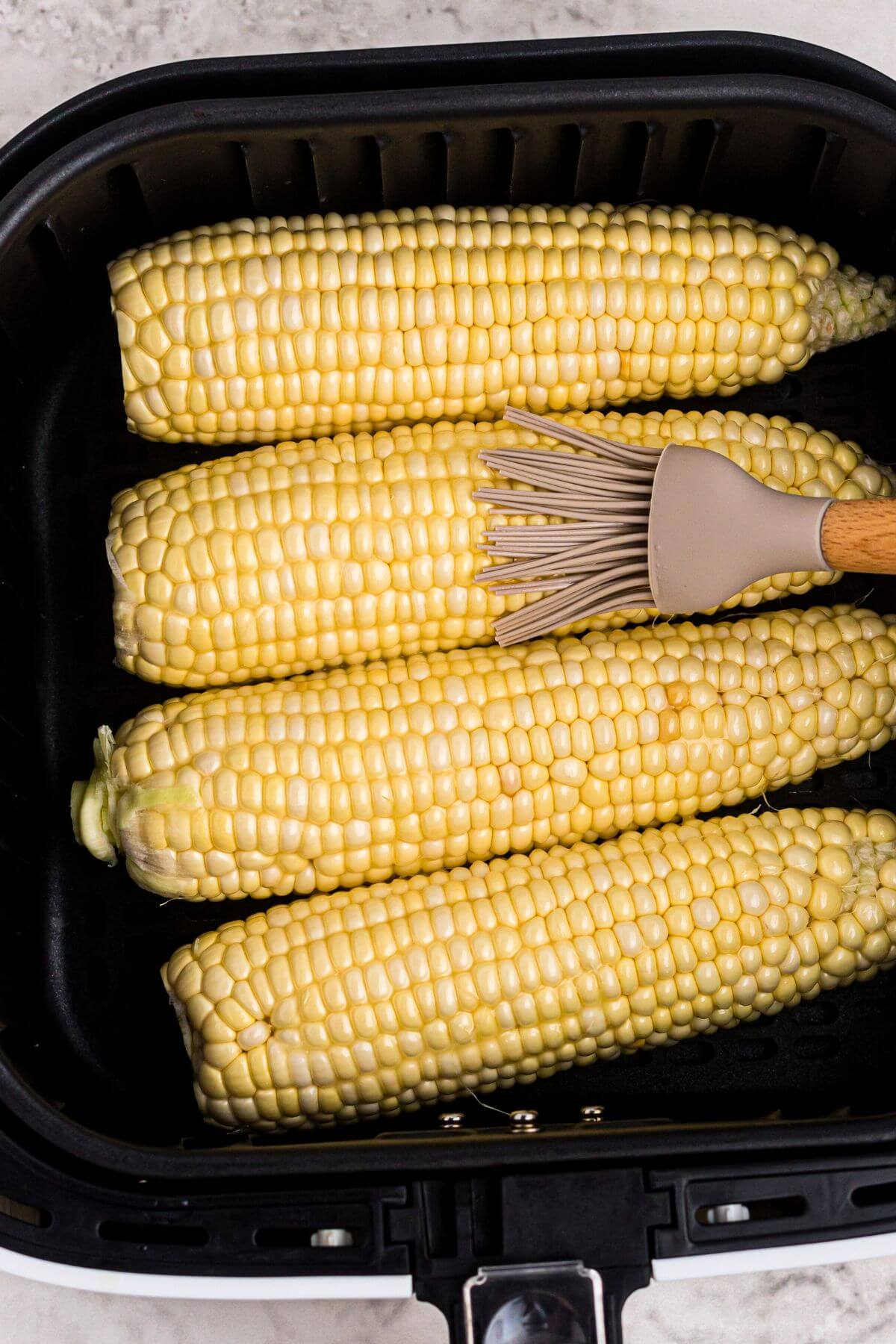Ears of corn in the air fryer basket, being brushed with melted butter before cooking. 