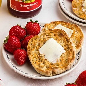 Golden crispy English muffins with a piece of butter on top, on a white plate served with strawberries and orange juice.