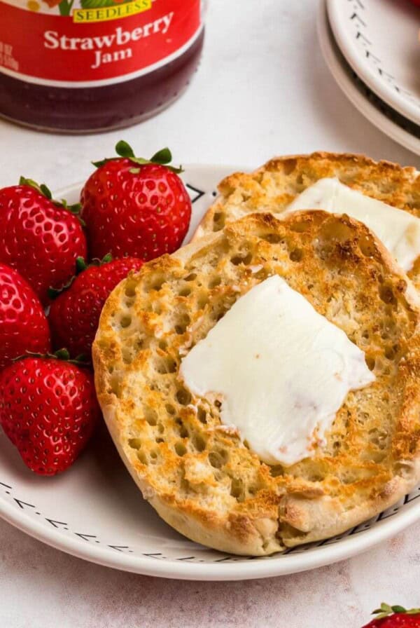 Golden crispy English muffins with a piece of butter on top, on a white plate served with strawberries and orange juice.