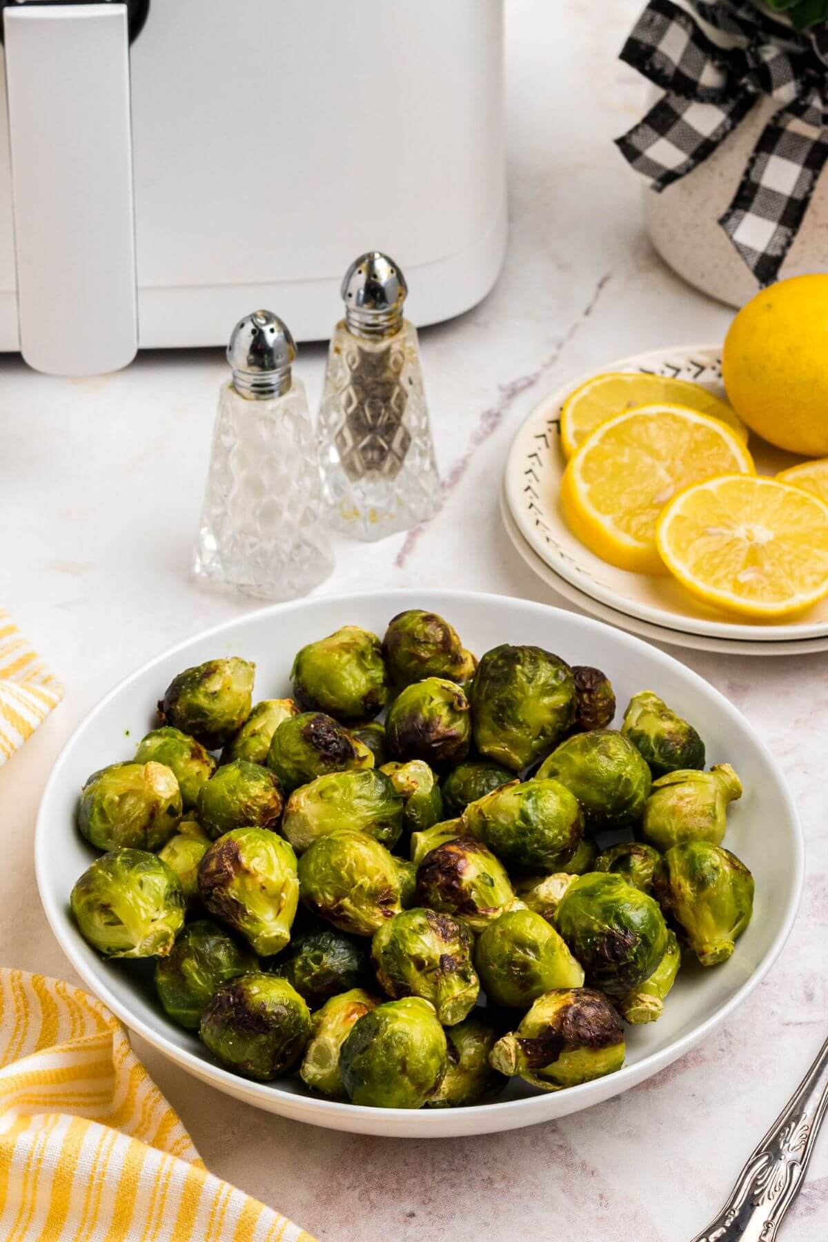 Green and juicy brussels sprouts in a white bowl in front of an air fryer with lemon slices on a plate. 