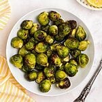 Green and juicy brussels sprouts in a white bowl in front of an air fryer with lemon slices on a plate.