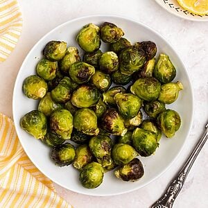 Green and juicy brussels sprouts in a white bowl in front of an air fryer with lemon slices on a plate.
