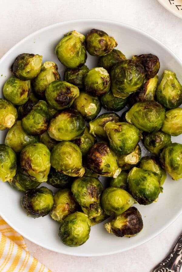 Green and juicy brussels sprouts in a white bowl in front of an air fryer with lemon slices on a plate.