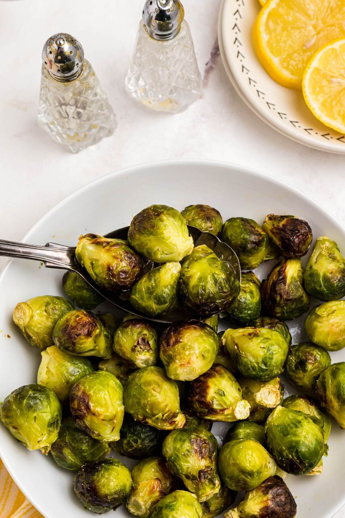 Green and juicy brussels sprouts in a white bowl in front of an air fryer with lemon slices on a plate and a spoon scooping out brussels sprouts. 