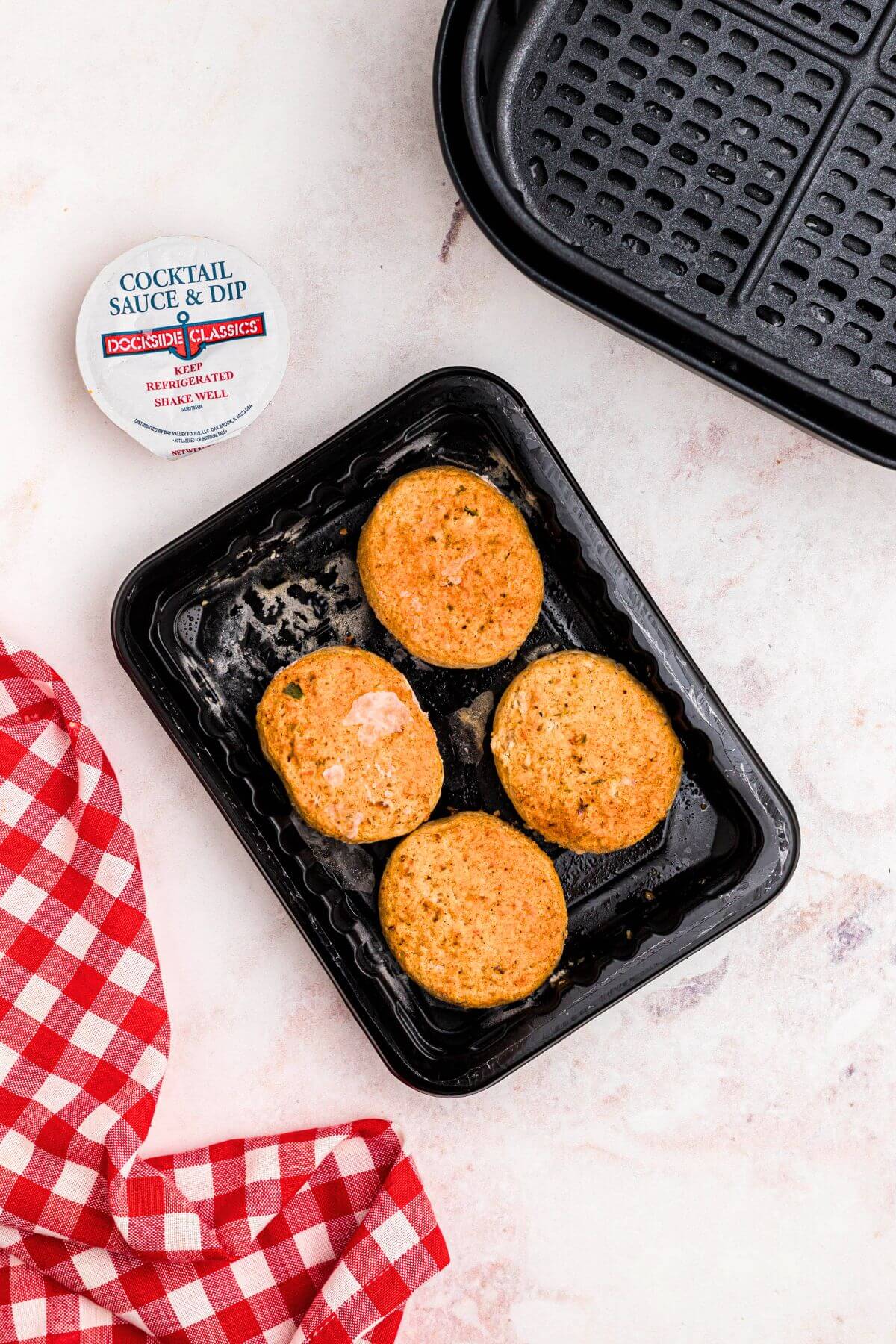 Frozen crab cakes on a black plastic plate in front of the air fryer basket. 