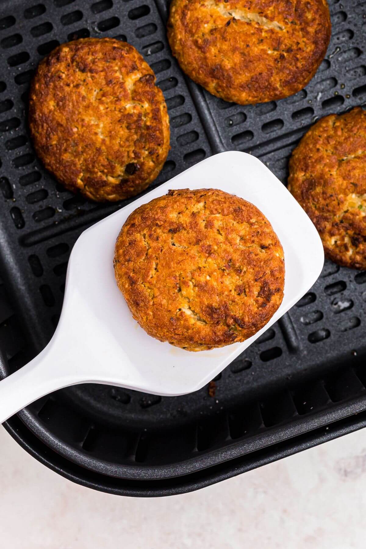 Golden crispy crab cakes in the air fryer basket with one being lifted by a white spatula. 