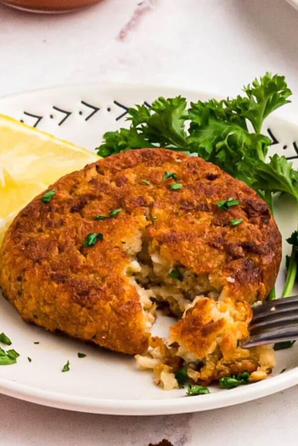 Golden crispy crab cake on a small white plate with a fork removing a bite and with a small bowl of cocktail sauce on the table for dipping.