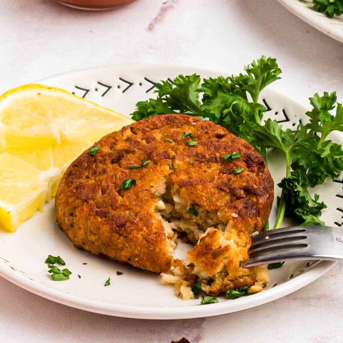 Golden crispy crab cake on a small white plate with a fork removing a bite and with a small bowl of cocktail sauce on the table for dipping.