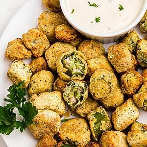 Golden breaded okra stacked on a white plate with a small dish or ranch dressing for dipping