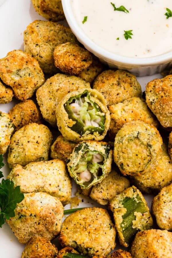 Golden breaded okra stacked on a white plate with a small dish or ranch dressing for dipping