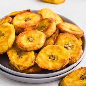Golden plantain slices on a small plate in front of the air fryer.