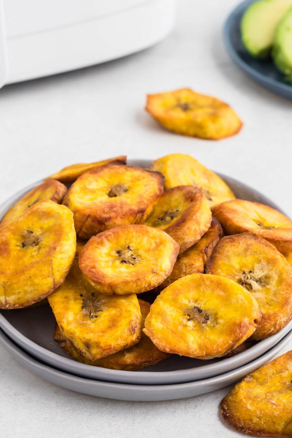 Golden plantain slices on a small plate in front of the air fryer. 