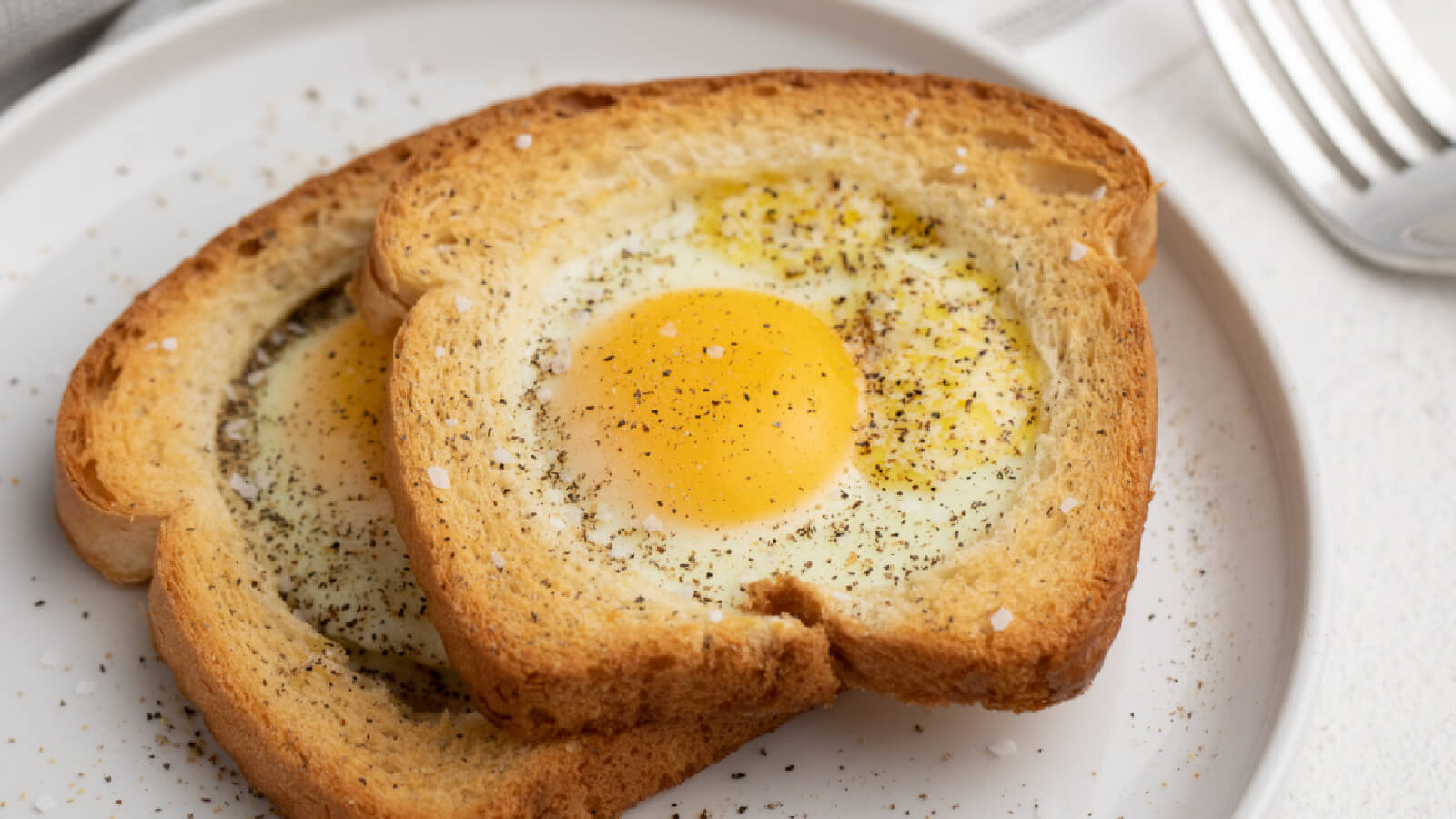 Egg in a basket made in the air fryer, garnish with salt and pepper and served on a white plate.