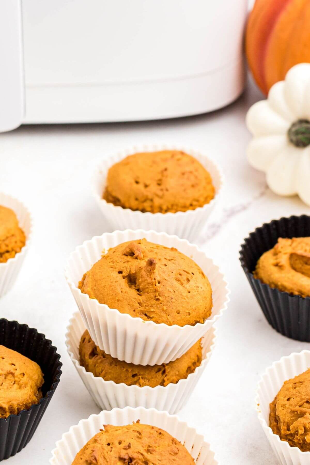 Pumpkin muffins scattered on a marble table in front of an air fryer, while still in their black or white silicone liners. 
