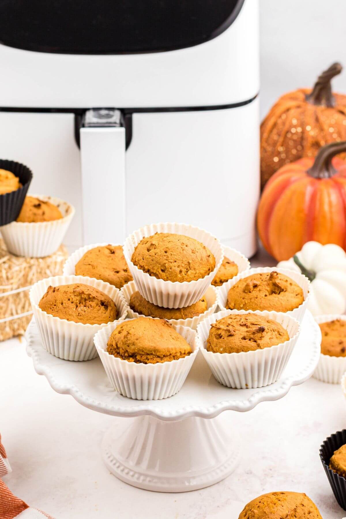 Golden orange pumpkin muffins on a cake stand, in front of the air fryer. 
