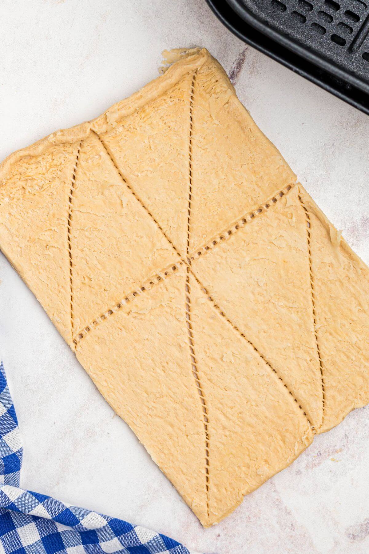A sheet of crescent roll dough rolled out and sliced into triangles, in front of the empty air fryer basket. 