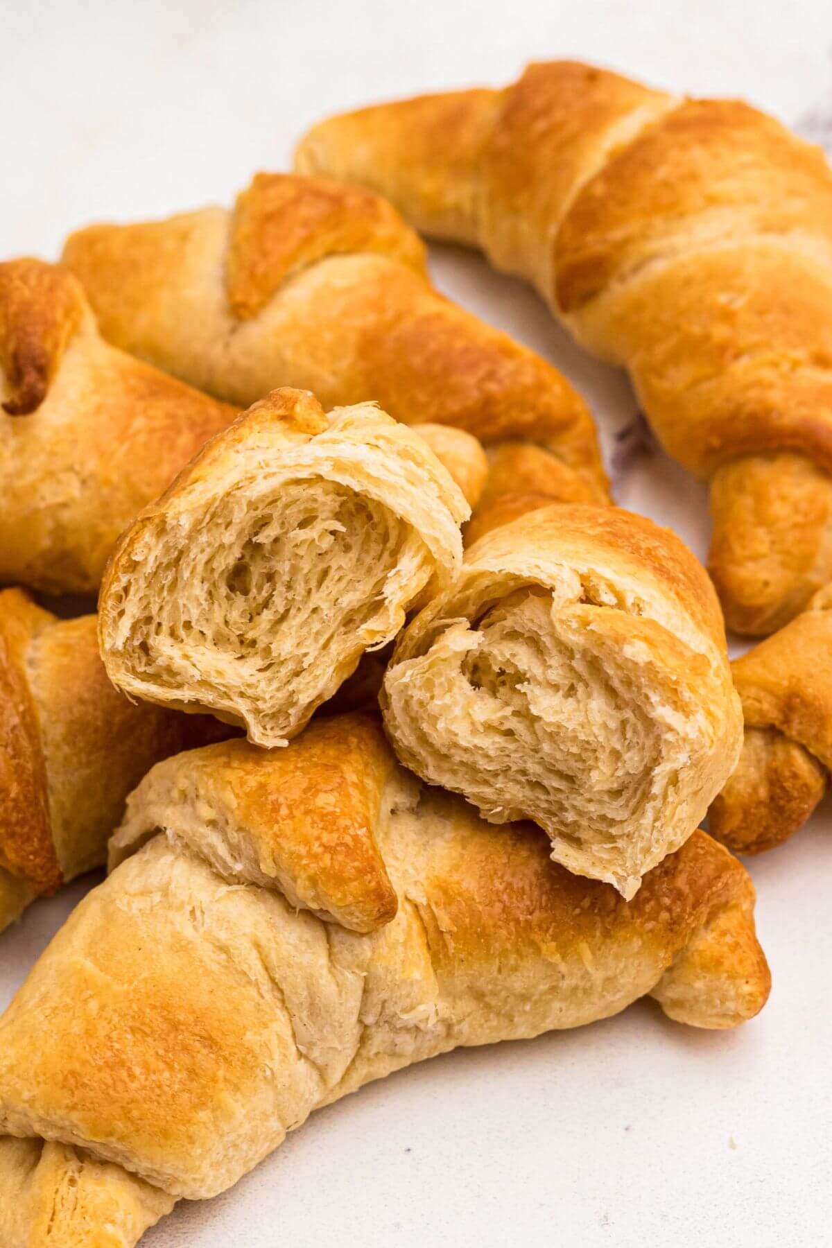 golden flaky crescent rolls on a white marble table with one split in two showing the flaky layers inside. 