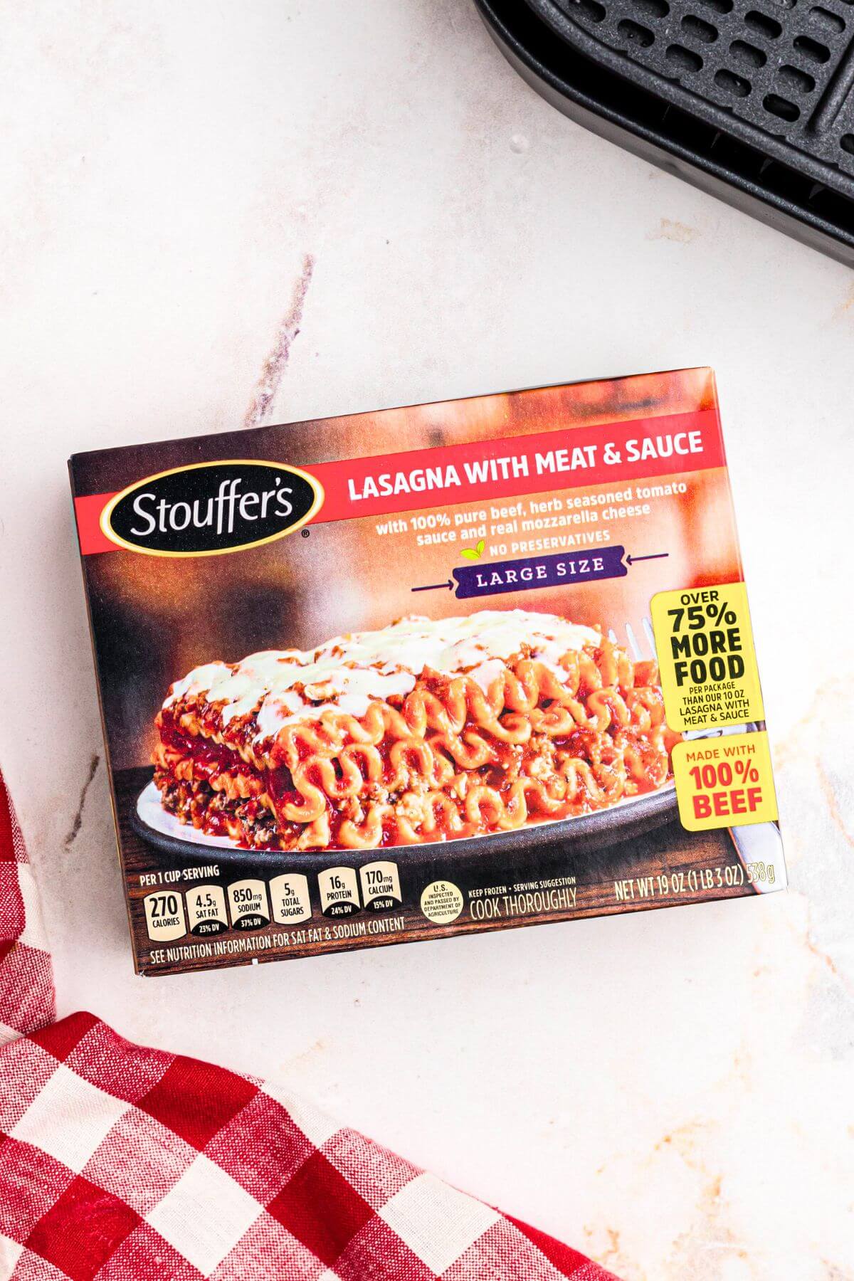 Box of frozen lasagna on a white marble table in front of the air fryer basket. 