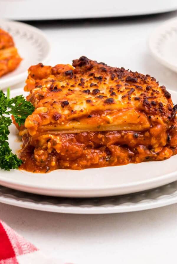 Cheesy cooked slice of lasagna with sauce on a white plate garnished with parsley.