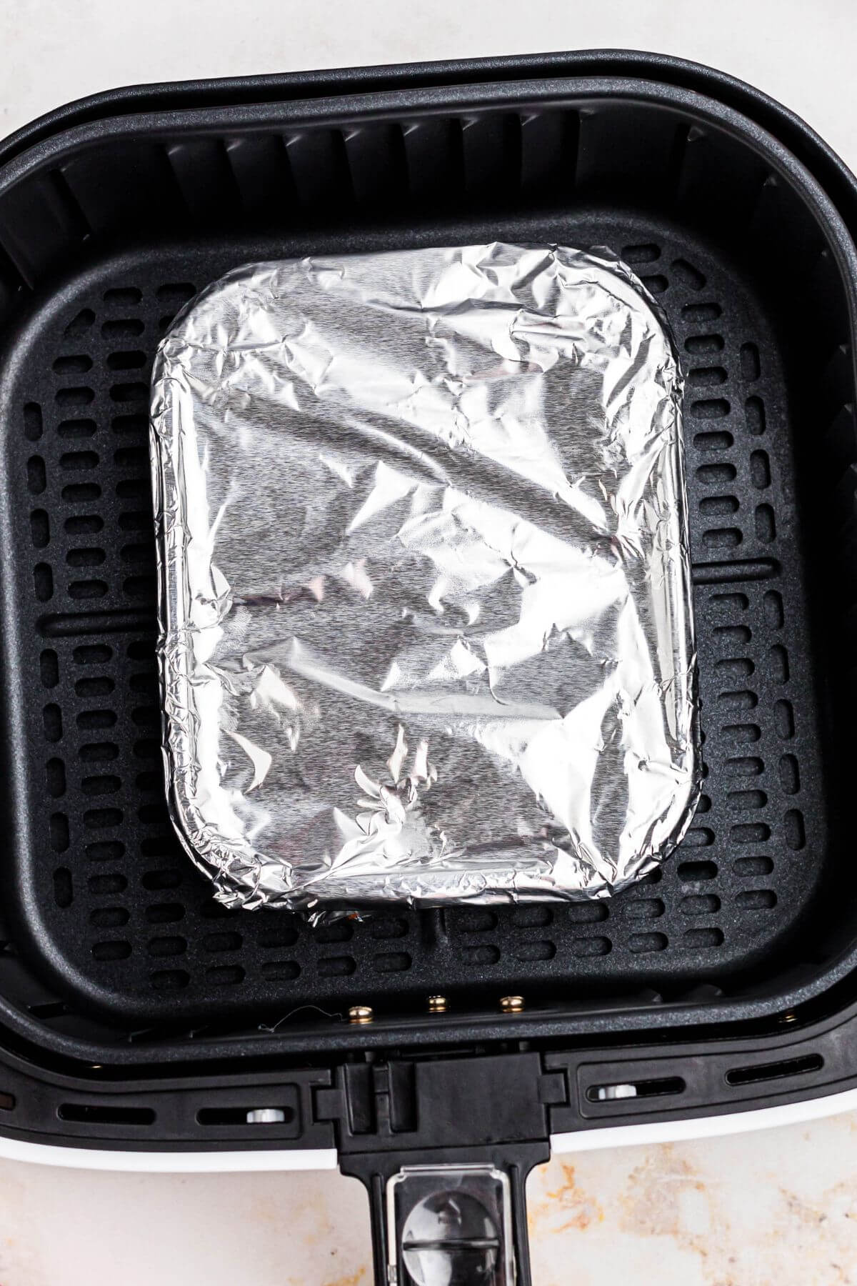 Frozen lasagna wrapped in a piece of aluminum foil, in the air fryer basket. 