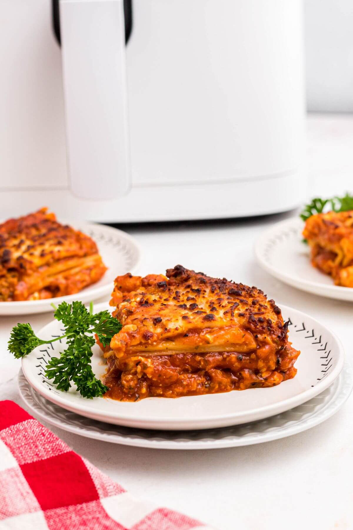 Cheesy cooked slice of lasagna with sauce on a white plate garnished with parsley.