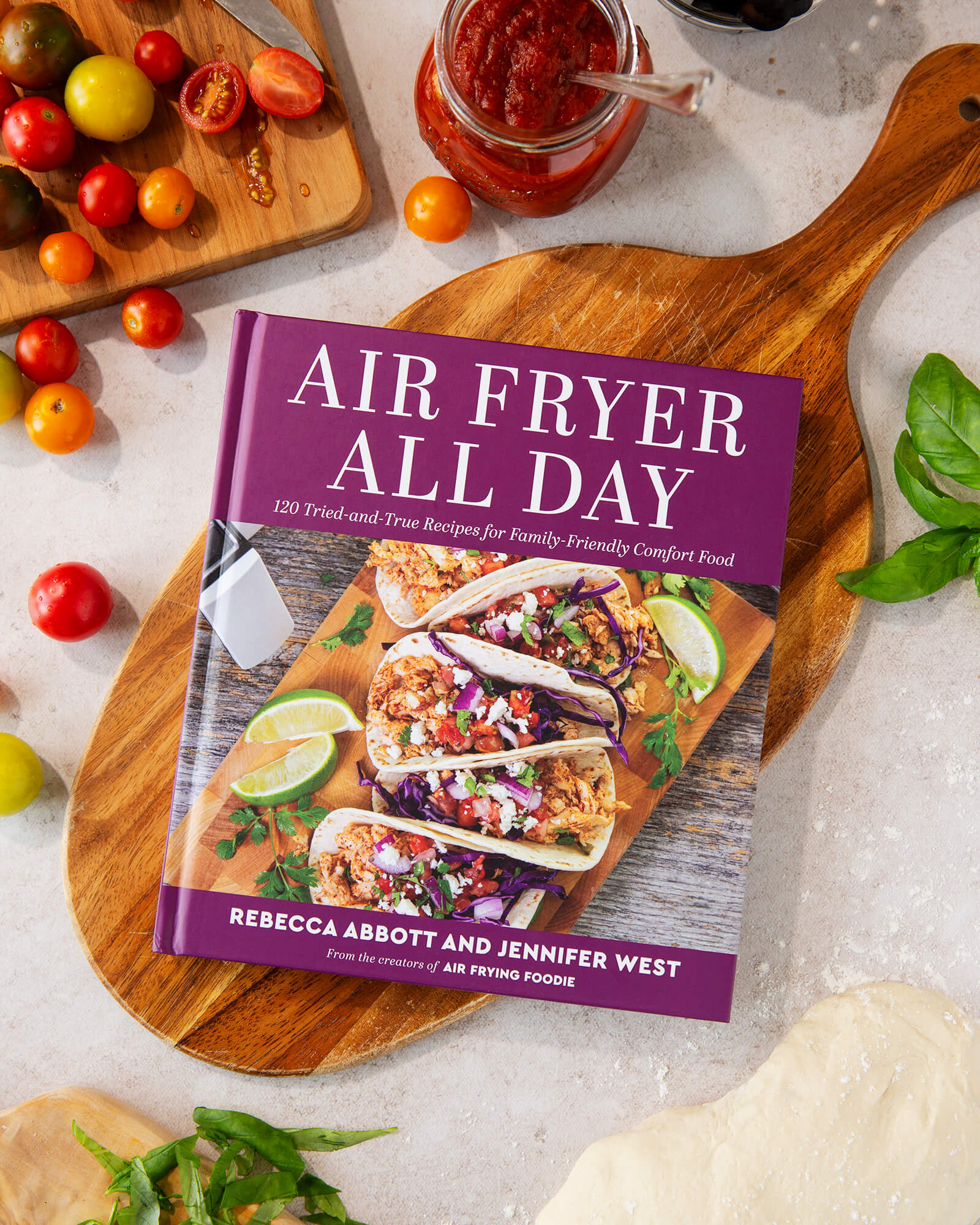 Photo of the air fryer all day cookbook on a cutting board with food surrounding it on a table.
