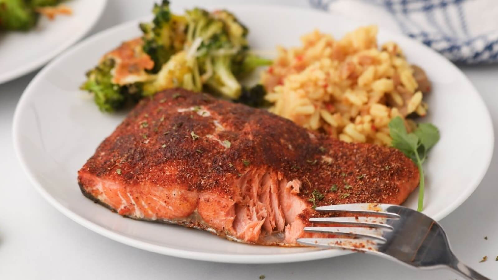 Simple Salmon Filet seasoned and sir fried, then served on a white plate with a side of brocolli and rice in the background, with a fork at the front of the plate.