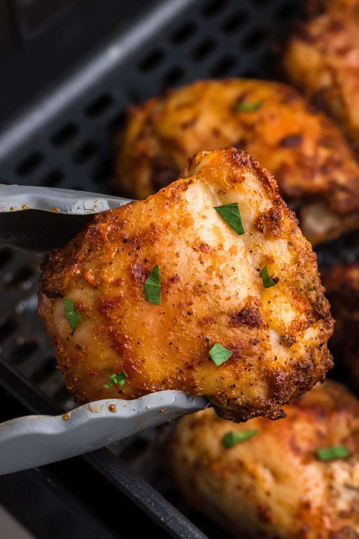 Crispy golden chicken thighs in the air fryer basket with one being lifted with tongs. 