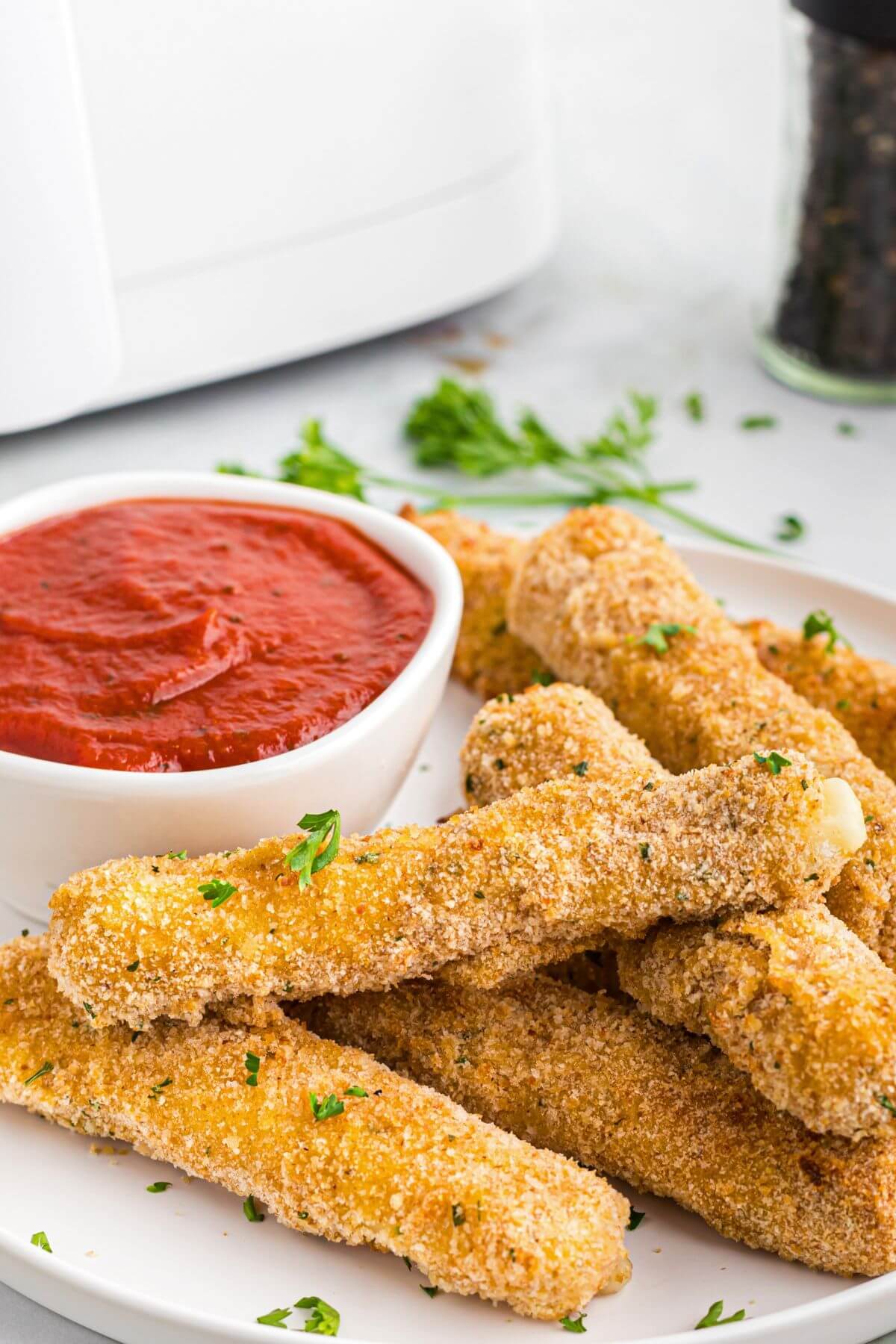 Golden crispy breaded cheese sticks stacked on a white plate with warm marinara sauce. 