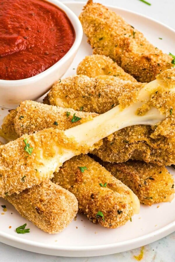 Golden breaded mozzarella cheese sticks stacked on a white plate with marinara sauce.