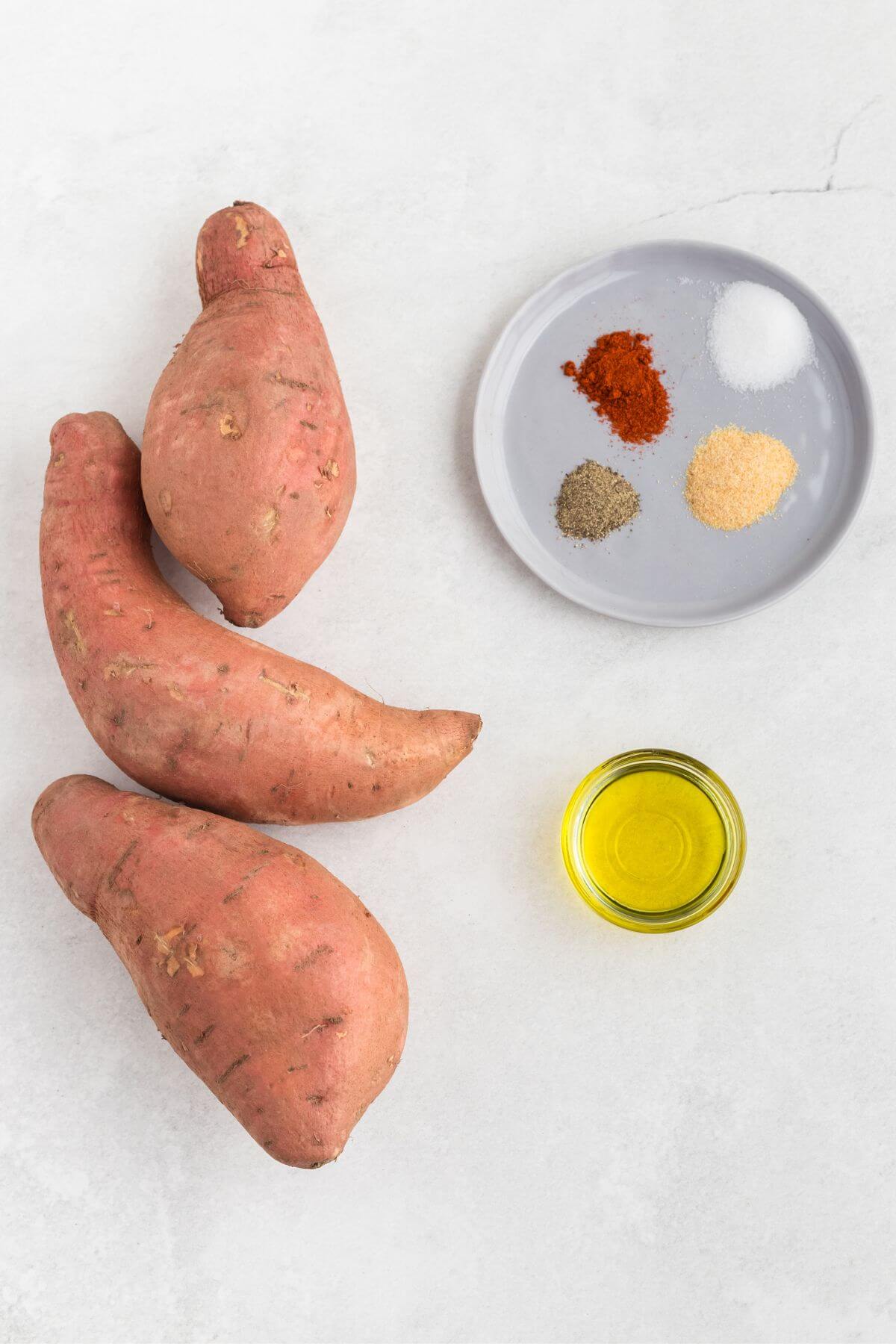 Sweet potatoes, olive oil, and seasonings on a white marble table. 