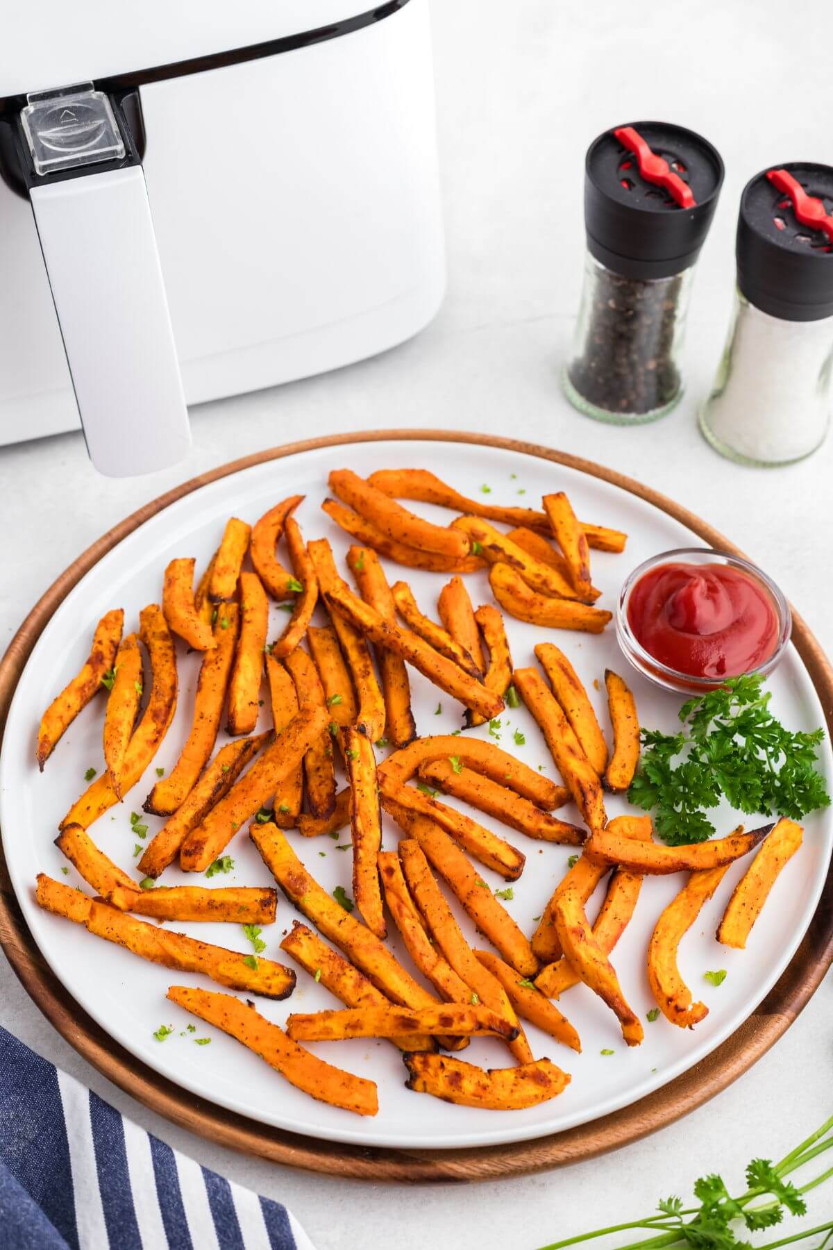 Orange golden sweet potato fries on a round white plate with a small dish of ketchup in front of a white air fryer. 