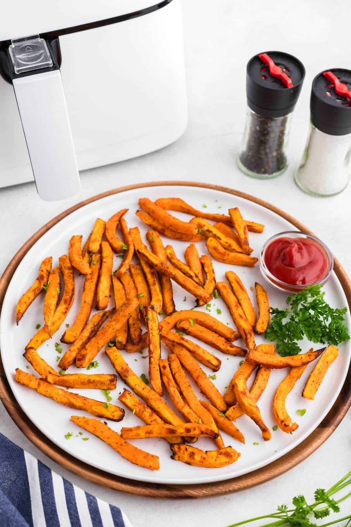 Orange golden sweet potato fries on a round white plate with a small dish of ketchup in front of a white air fryer. 