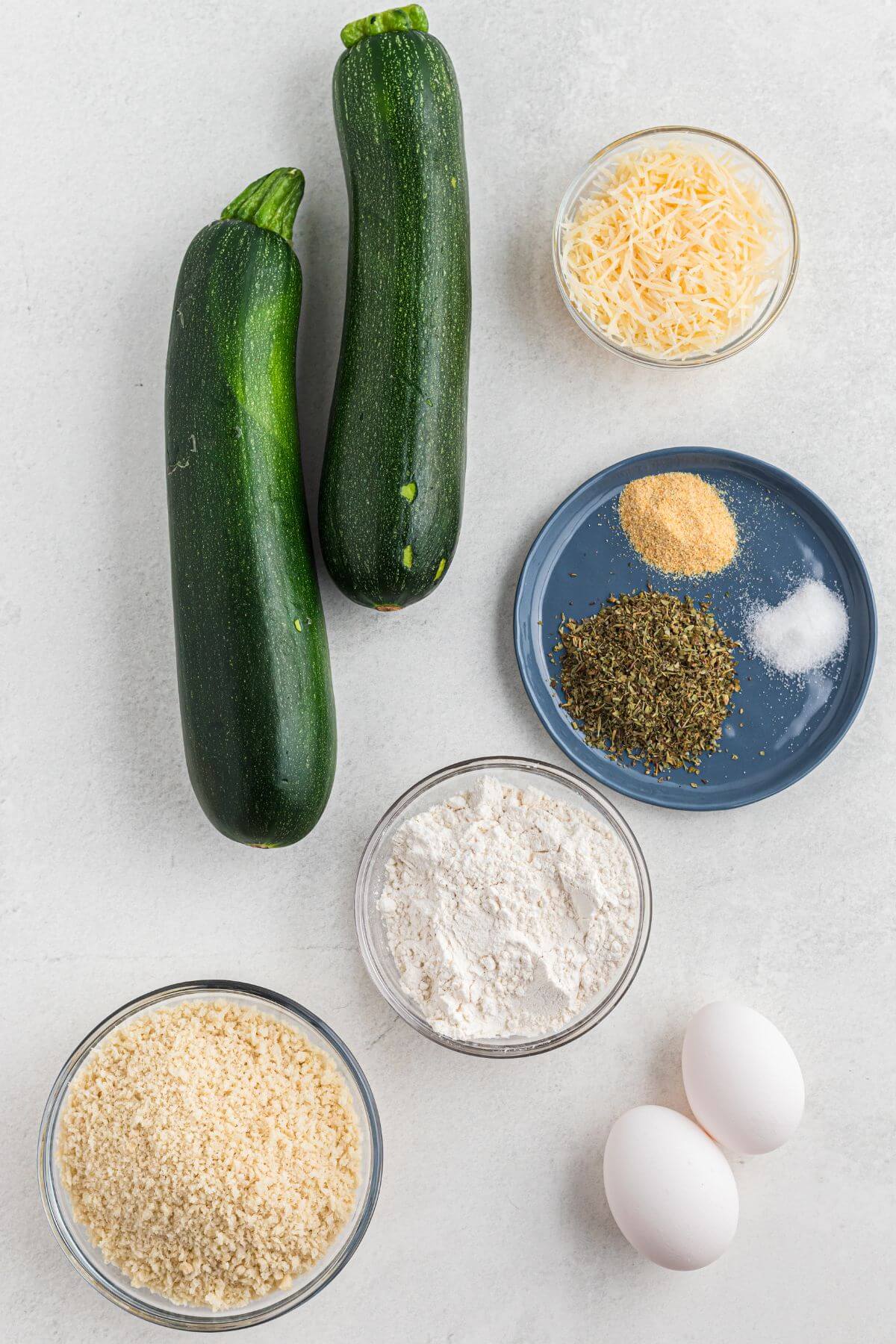 Zucchini, breadcrumbs, parmesan cheese, flour, eggs, and seasonings measured on a white marble table. 